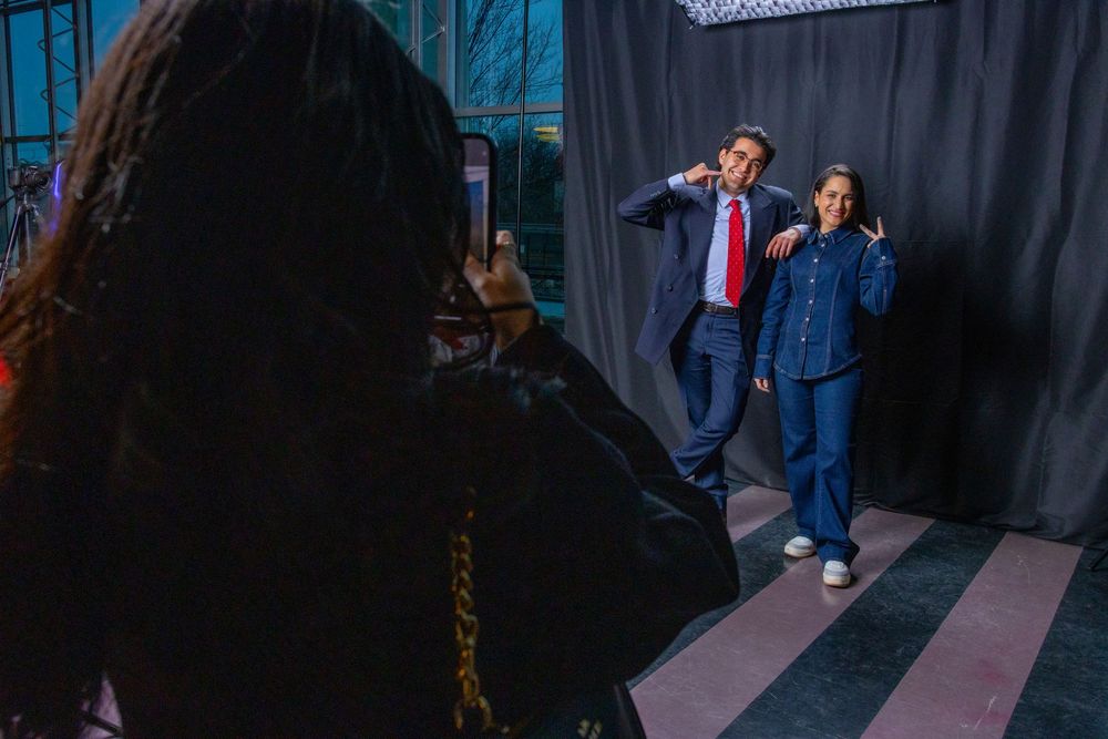 Two people pose playfully in front of a gray backdrop while a photographer captures their photo indoors.