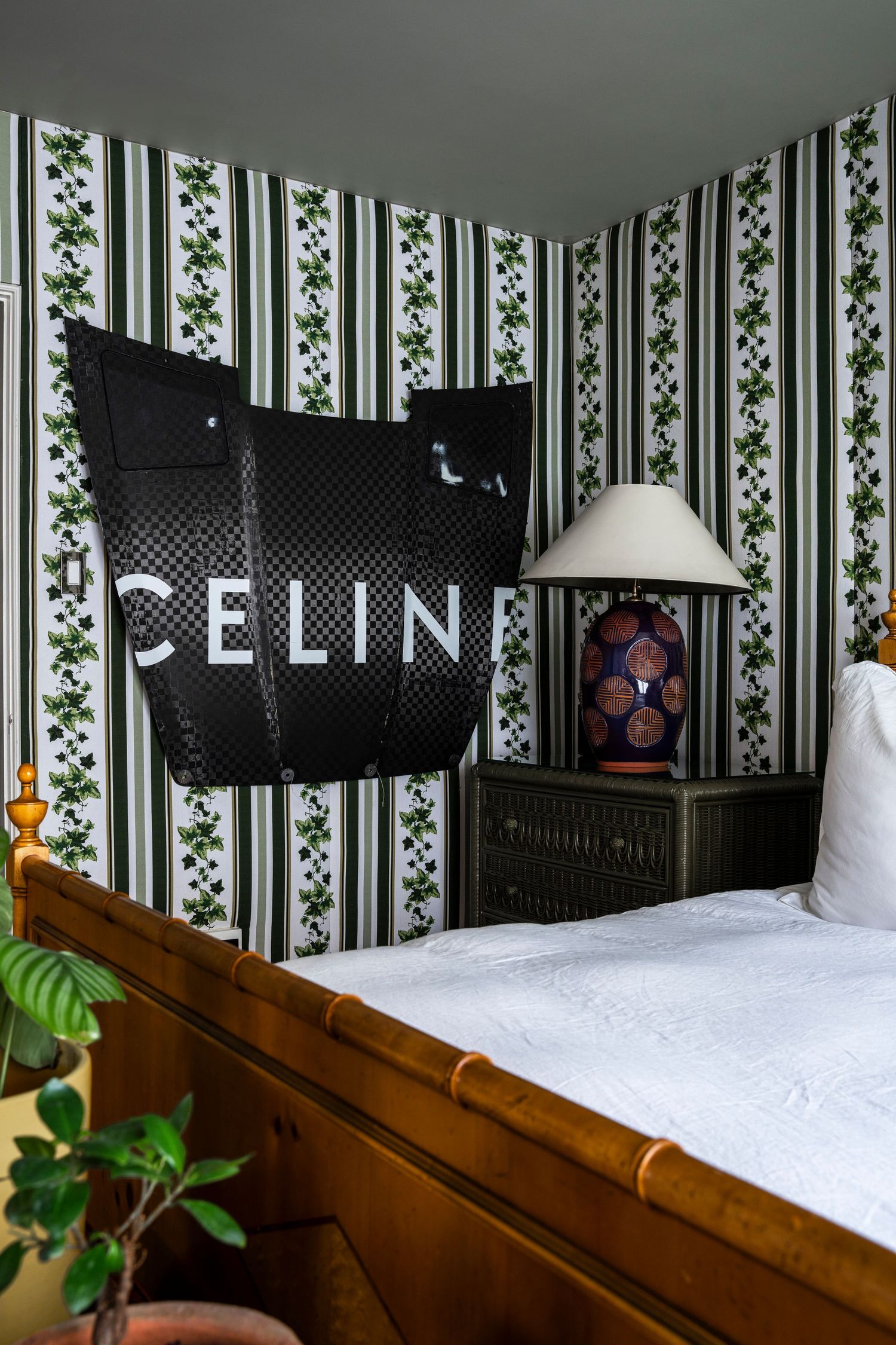 Bedroom with floral striped wallpaper, bamboo bed frame, green nightstand, lamp, and a black Celine bag displayed on the wall.