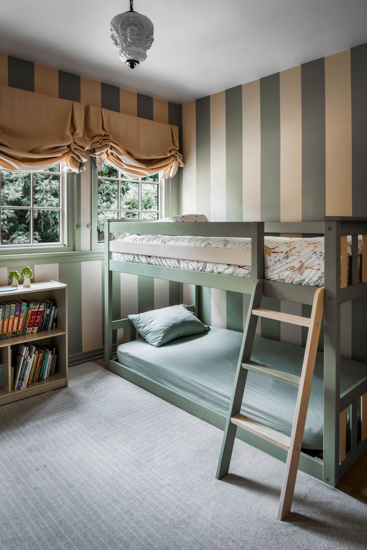 Sage green bunk beds with ladder in striped wallpaper kids room featuring Roman shade window and built-in bookshelf.