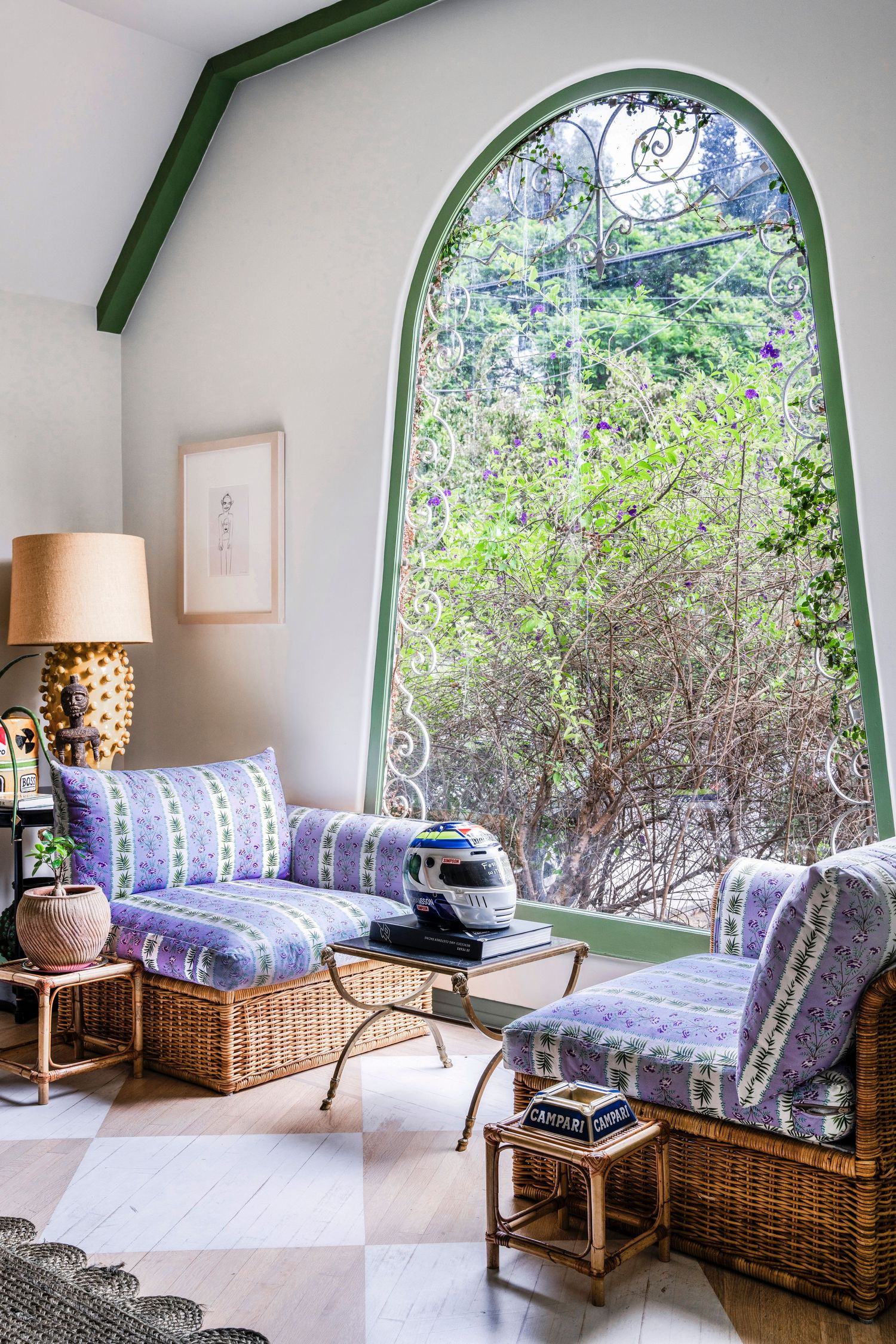 Cozy sitting area with wicker chairs, purple upholstery, and a large arched green-framed window overlooking lush greenery.