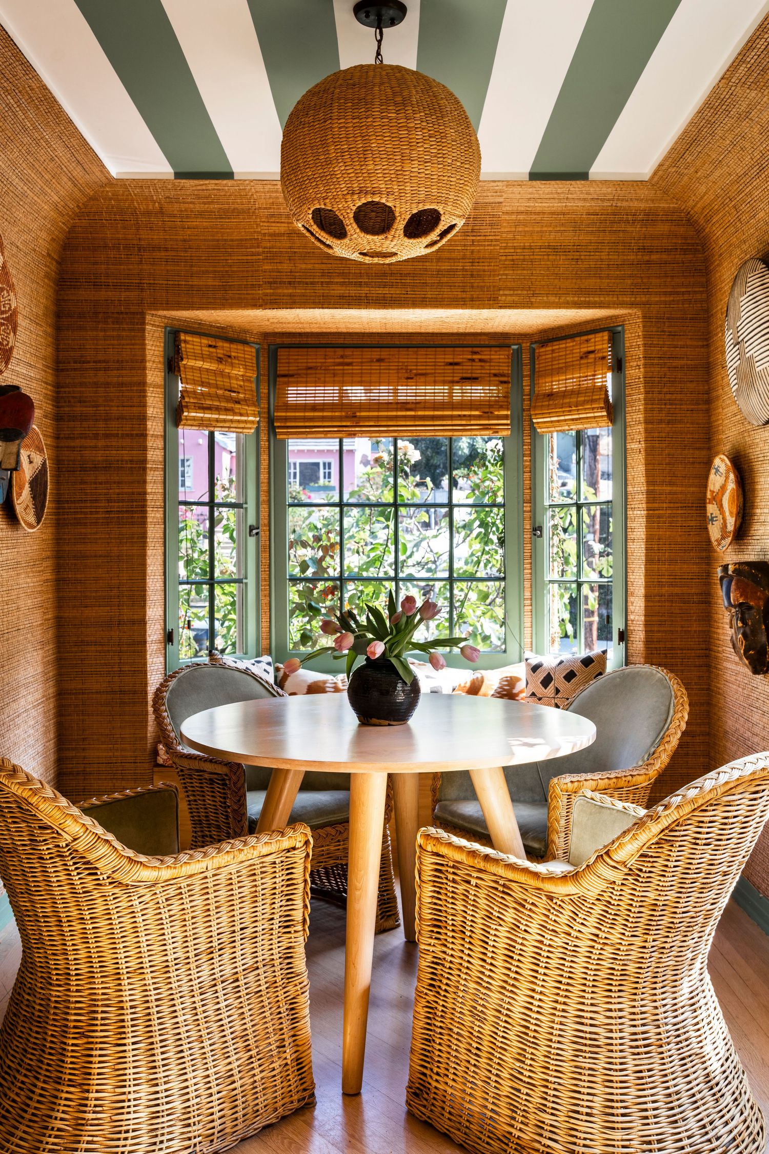 Cozy dining nook with wicker chairs, round wood table, bamboo shades, grasscloth walls, and striped green ceiling with rattan pendant light.