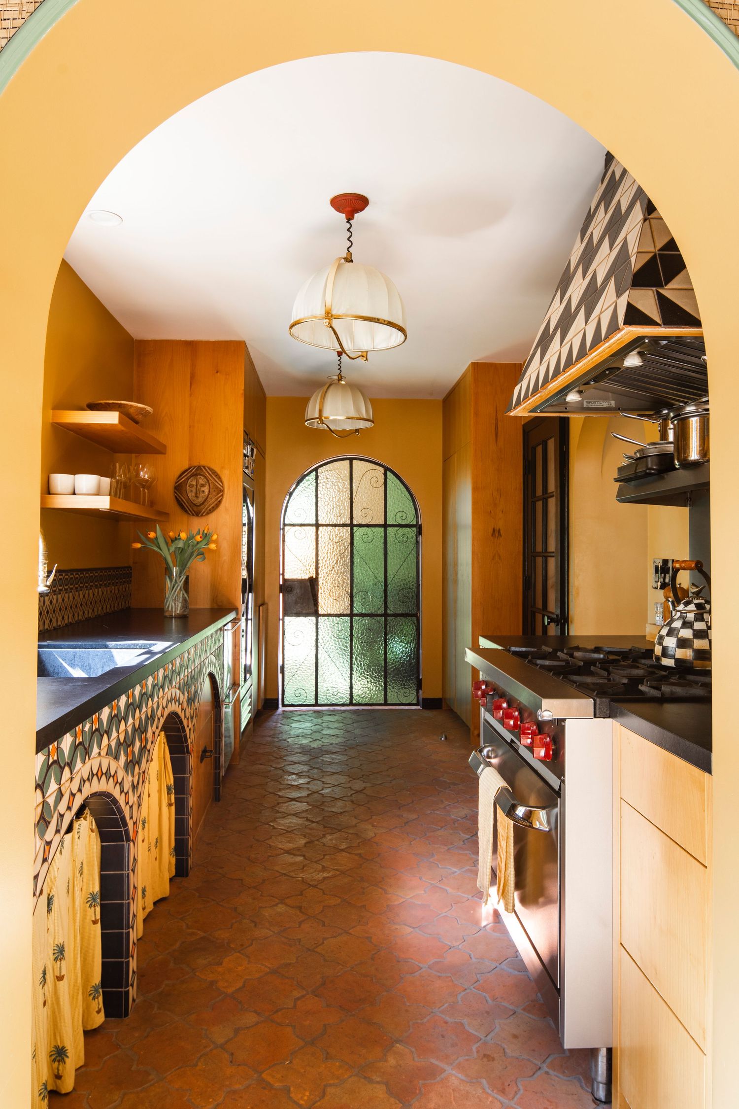 Warm yellow Mediterranean kitchen with terracotta tile floors, arched doorways, decorative tilework, pendant lights, and black countertops.