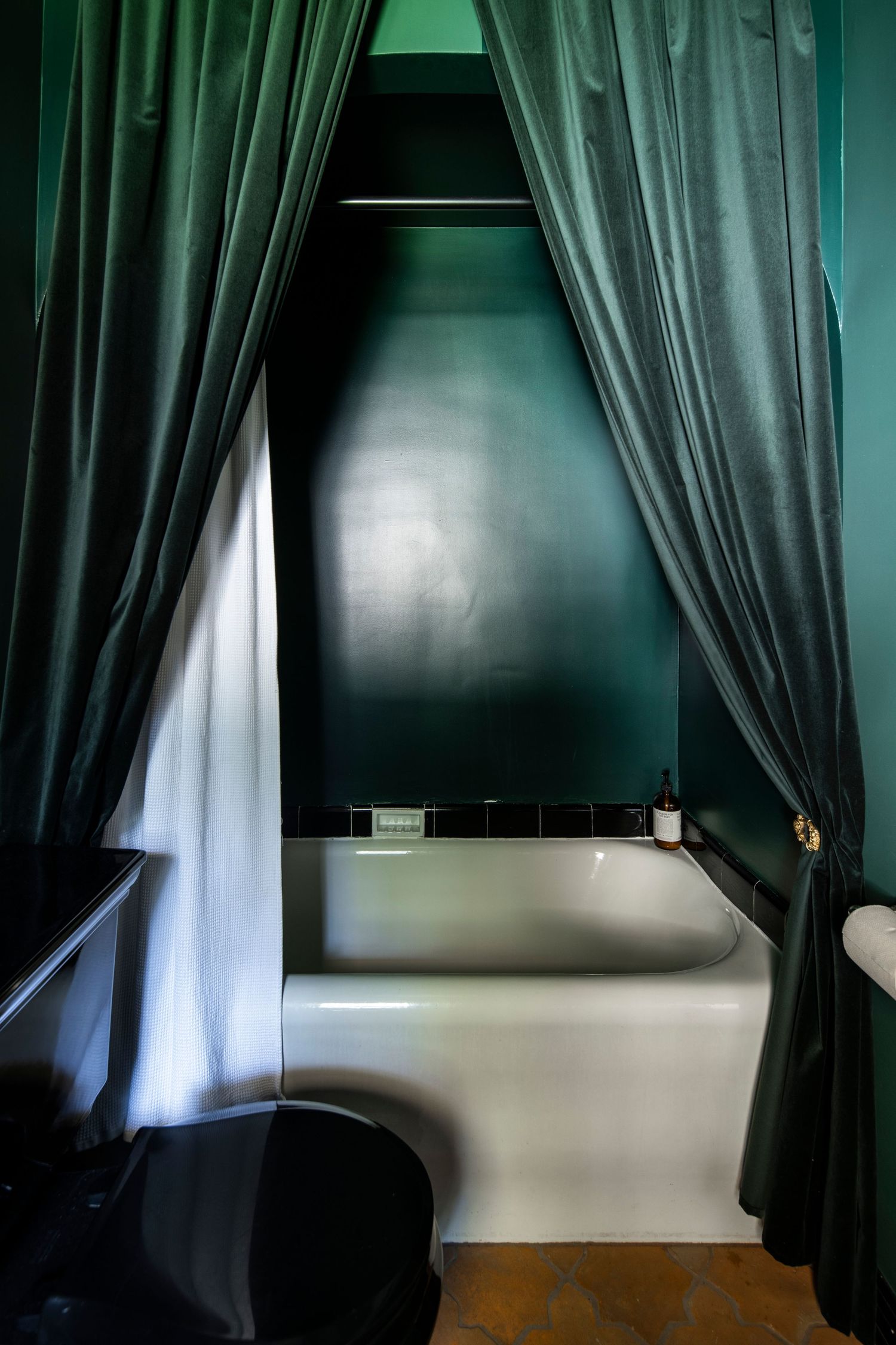 Dramatic dark green bathroom with velvet curtains framing a white soaking tub against a glossy emerald wall.