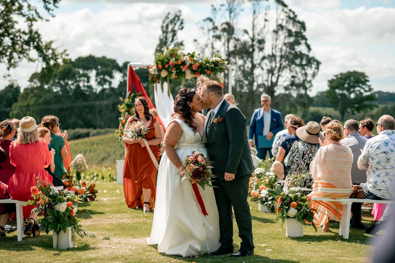 Bride and groom share first kiss at outdoor wedding ceremony with floral arch and guests seated on green lawn.
