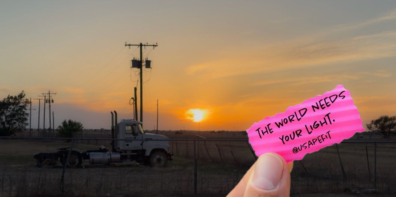 Hand holding pink note saying 'The world needs your light' against a sunset sky with silhouetted truck and tower.