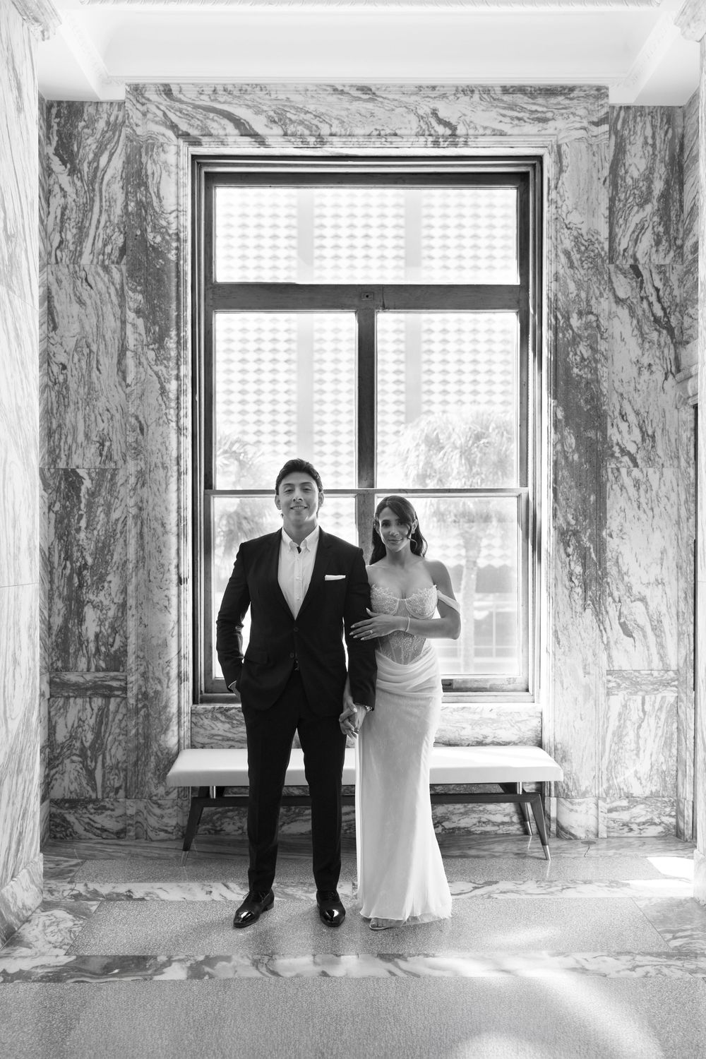 Black and white wedding portrait of bride and groom standing before a large window in an elegant marble-walled venue.