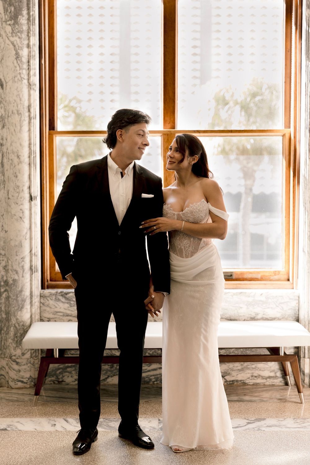 Bride and groom stand by a sunlit window, she in a white gown, he in a black suit, smiling at each other.