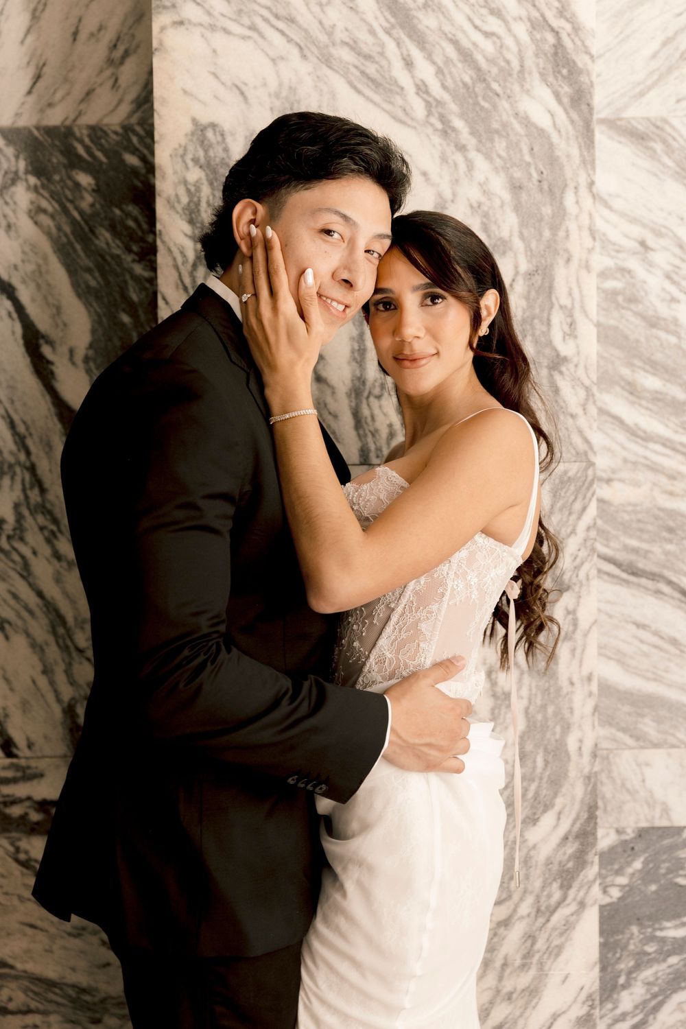 A bride and groom embrace tenderly in front of marble columns, she in a white lace gown and he in a black suit.