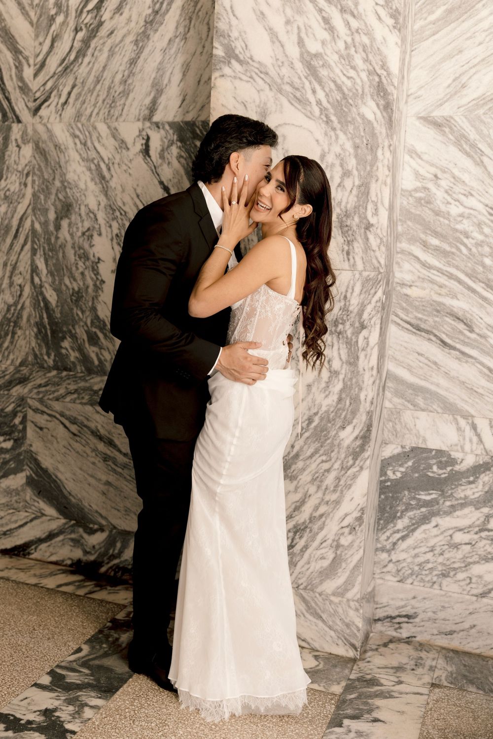 Bride in white lace gown and groom in black suit share a romantic kiss against a marble wall backdrop.