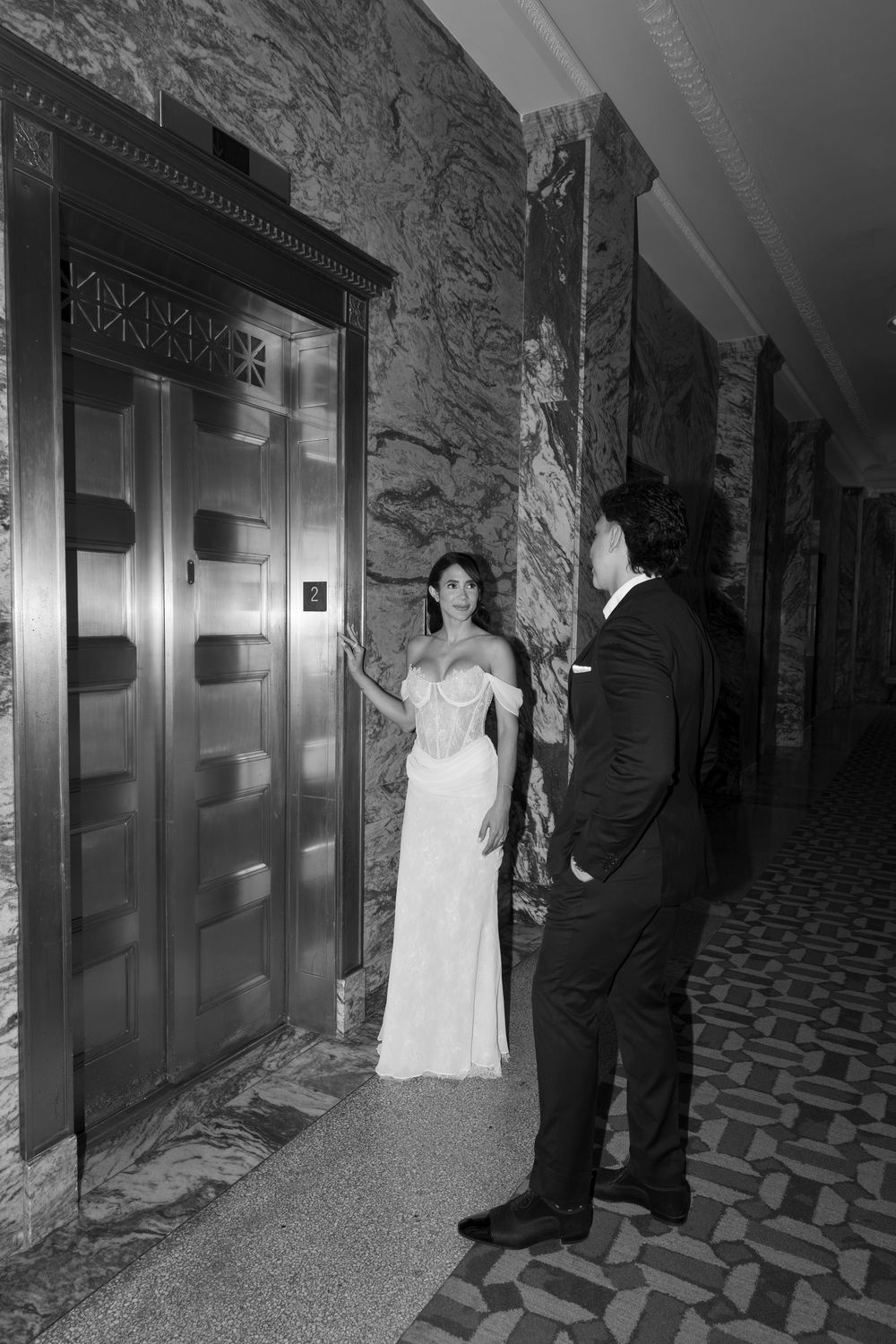 Black and white photo of bride in white gown and groom in tuxedo waiting by ornate elevator doors.