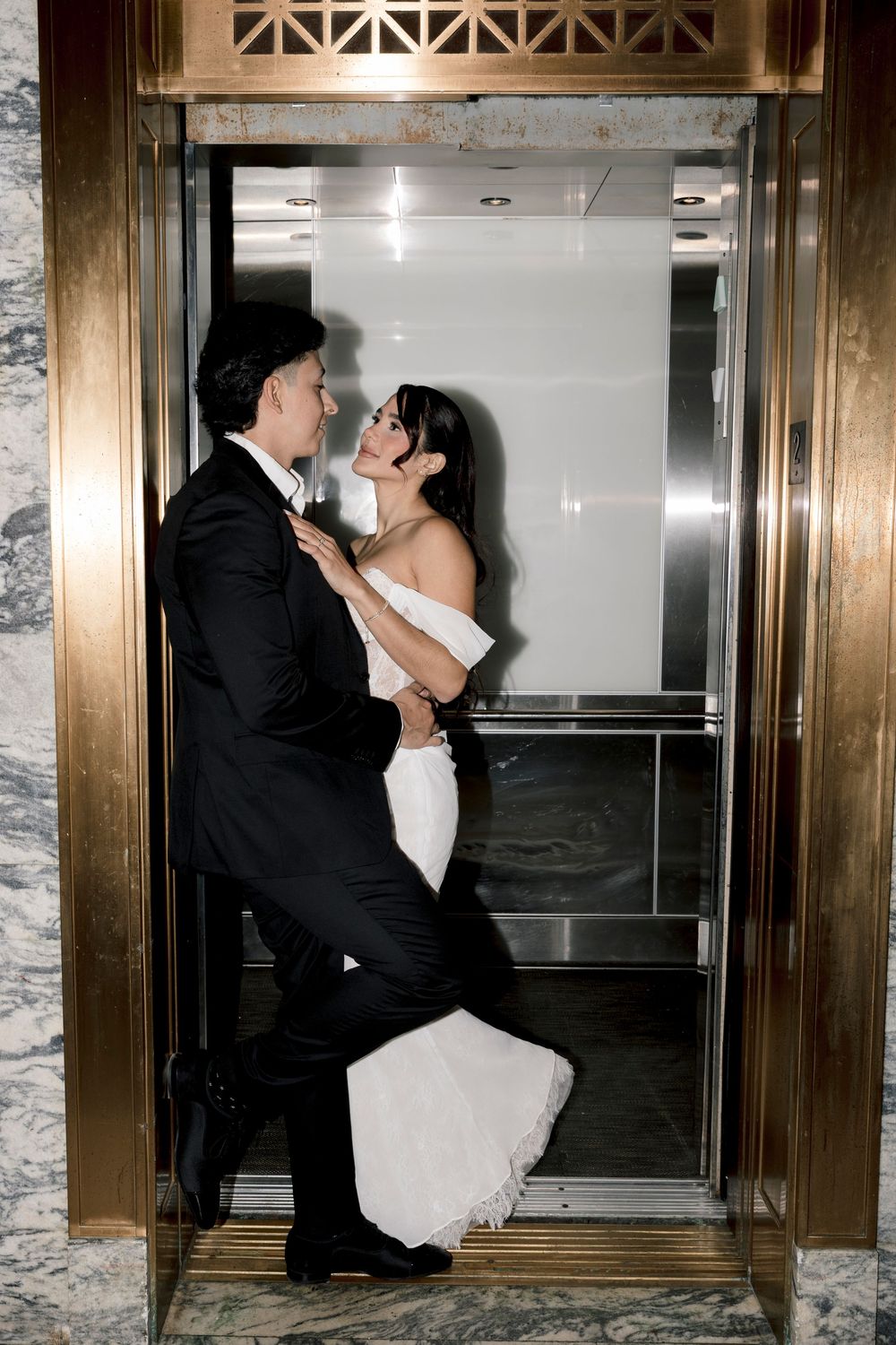 Bride and groom sharing a romantic moment in a gold elevator, bride in white gown, groom in black tuxedo.