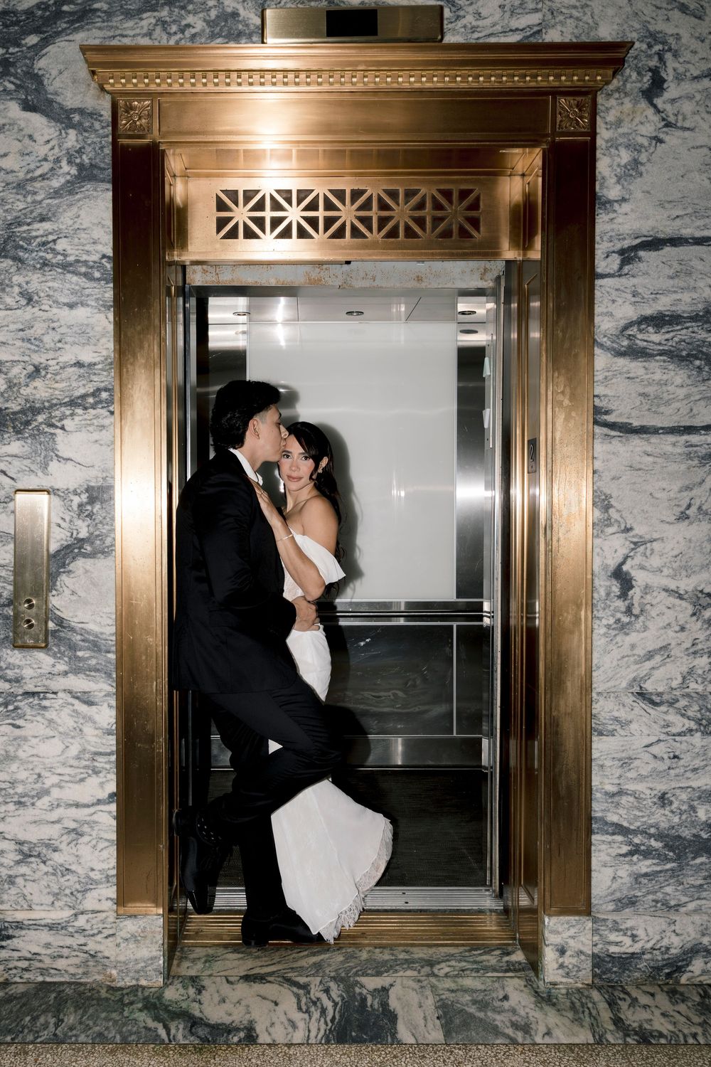 Bride and groom sharing a romantic kiss inside an ornate gold elevator with marble walls.