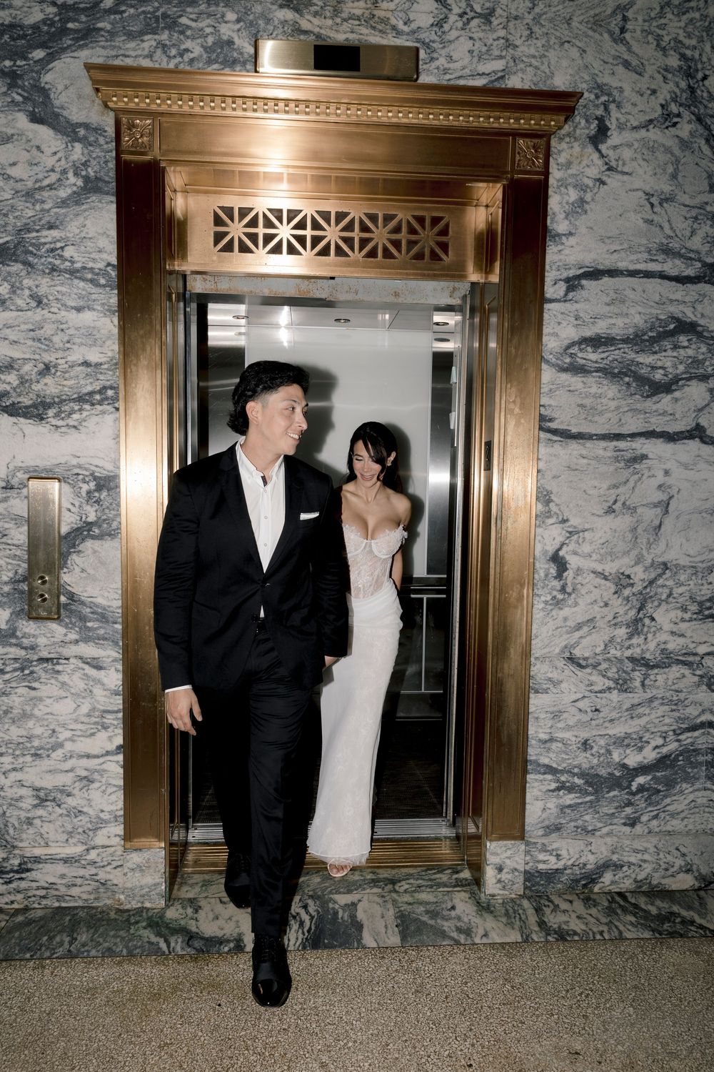 Bride in white gown and groom in black suit exiting a gold ornate elevator in a marble-walled venue.