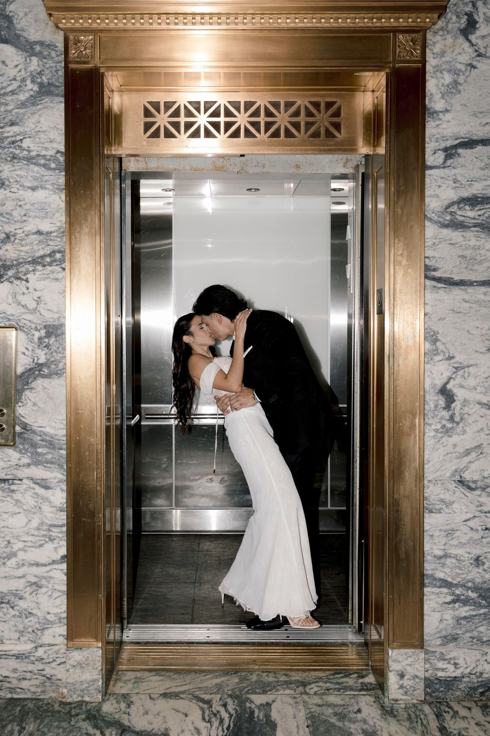 Newlyweds kissing passionately in a luxurious gold-framed elevator with marble surroundings.