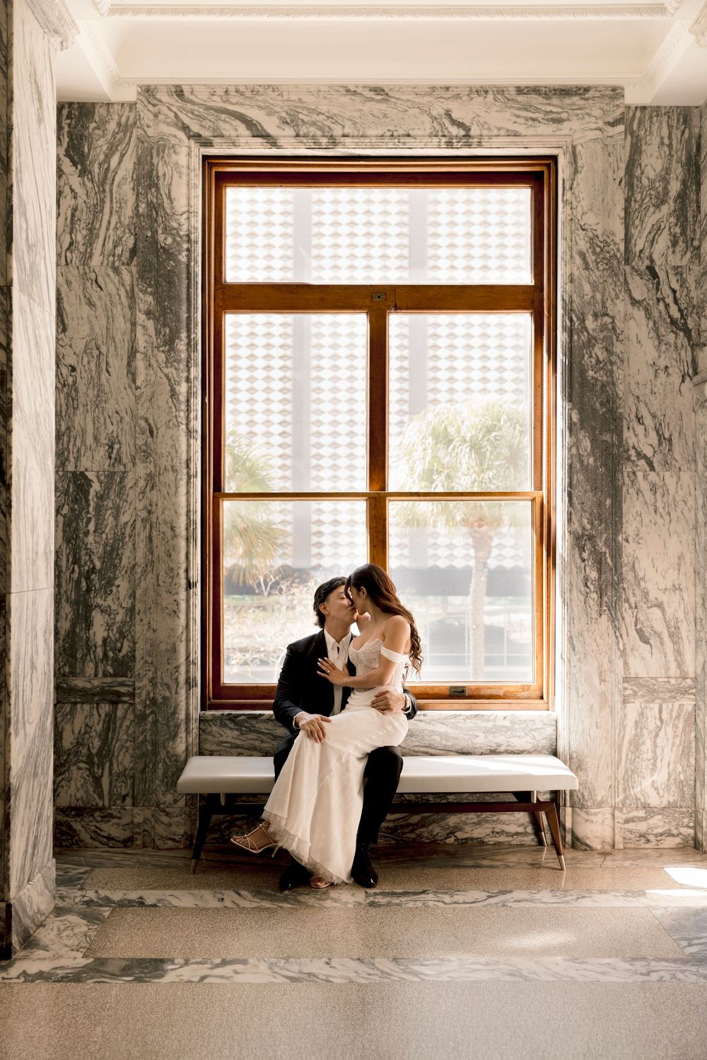 Bride and groom share a romantic kiss on a bench before a large window in an elegant marble-walled venue.