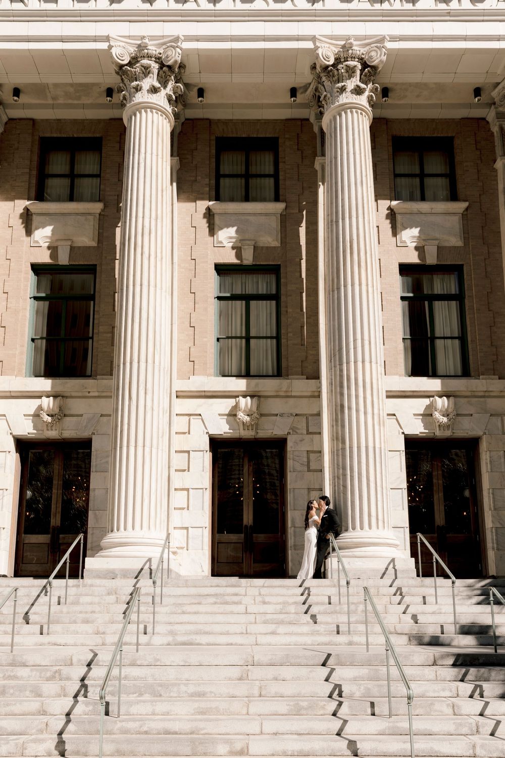 Bride and groom embrace on grand stone steps of a neoclassical building with tall Corinthian columns.