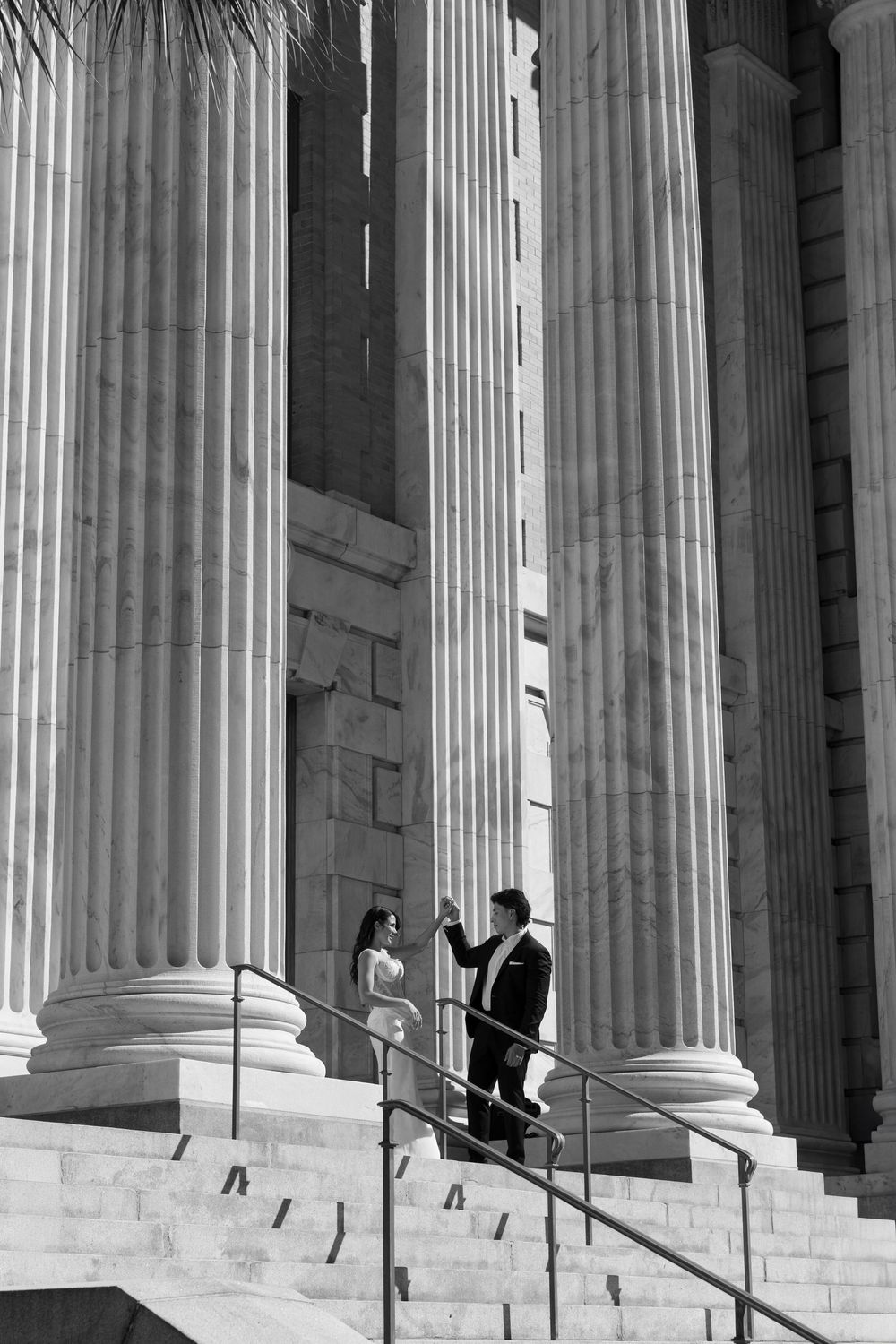 Black and white photo of a couple in formal attire on steps between grand classical columns.