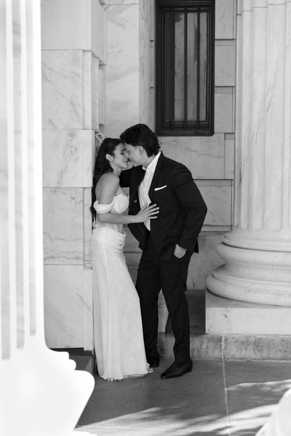 Black and white photo of a couple kissing in formal wedding attire between marble columns of an elegant building.