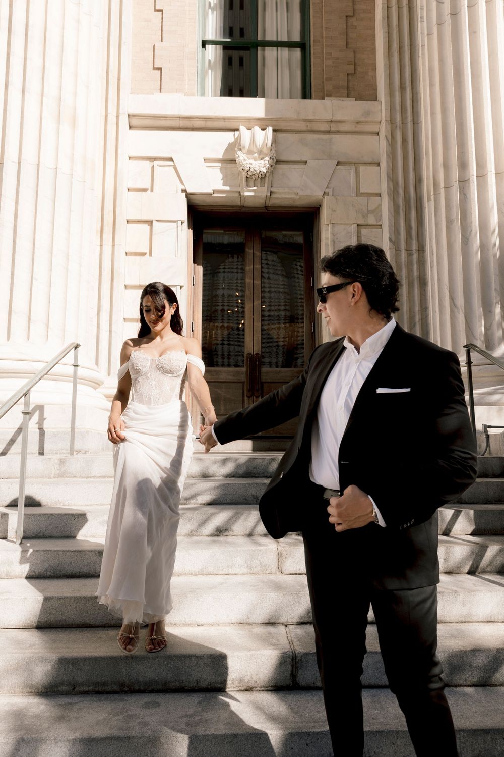 Elegant bride in white gown and groom in black suit holding hands on grand stone steps.
