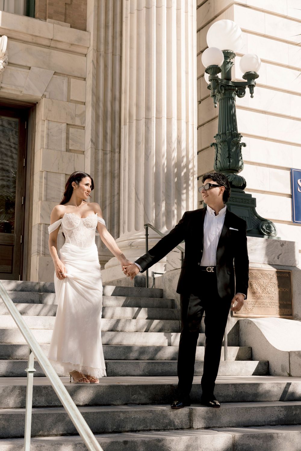 Stylish wedding couple holding hands descending sunlit stone steps of a classical building.