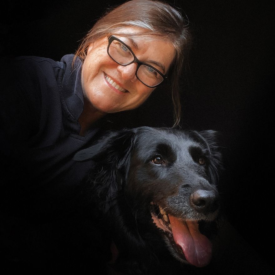 A smiling woman with glasses poses with a happy black dog against a dark background.