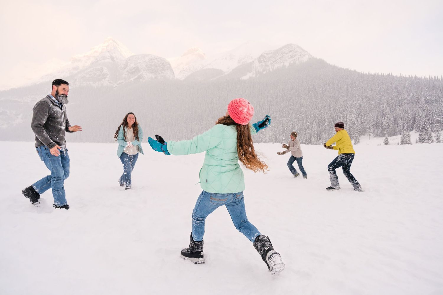Family playing snowball fight in snowy mountain landscape with snow-covered trees and pink sky.