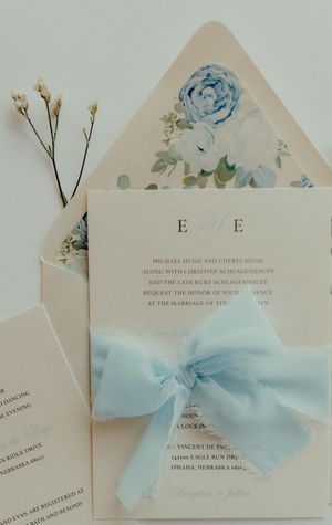 Elegant wedding invitation suite with light blue ribbon bow, floral envelope liner, and delicate white flowers on a soft white background.