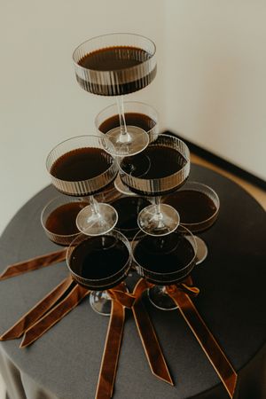Elegant pyramid tower of dark cocktails in coupe glasses with brown satin ribbons on a black round table.