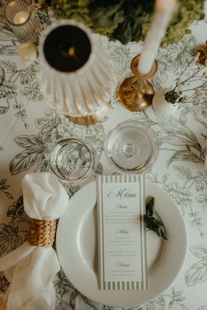 Elegant wedding table setting with floral tablecloth, striped menu card, white plate, napkin ring, and glassware.