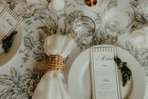 Elegant wedding table setting with floral tablecloth, white plates, gold napkin ring, and a menu card on a decorated tablescape.