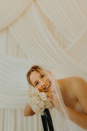 Smiling bride in white veil holding hydrangea bouquet against elegant draped fabric backdrop.