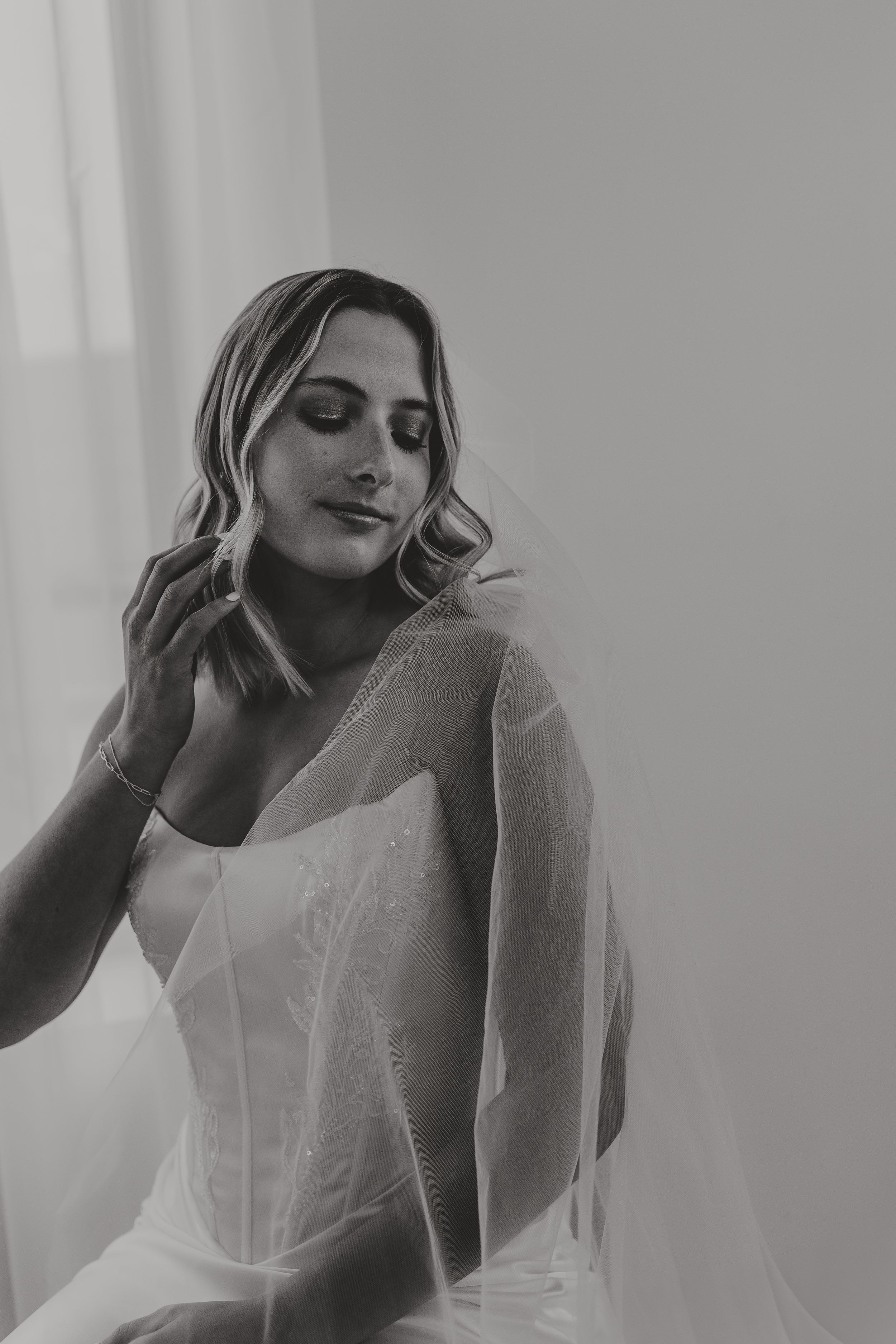 Black and white bridal portrait of a bride in a lace wedding gown and veil, posing gracefully near a window.