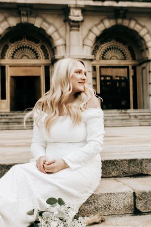 Blonde woman in white off-shoulder gown sits on stone steps before ornate building facade with arched windows.