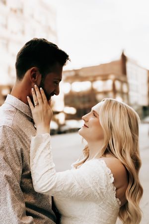 A blonde bride tenderly touches her groom's face while gazing into his eyes during a romantic outdoor engagement session.