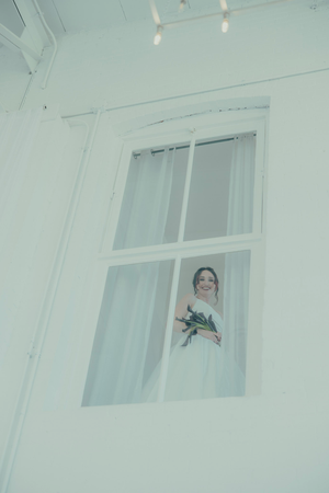 Smiling bride in white dress holding bouquet, viewed through large white-framed window in bright white room.