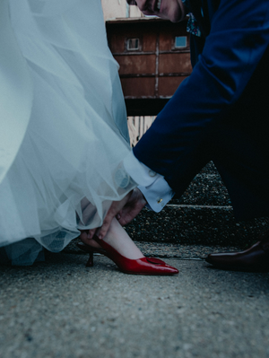 A groom in a blue suit kneels to adjust a bride's red kitten heel shoe beneath her flowing white wedding gown.