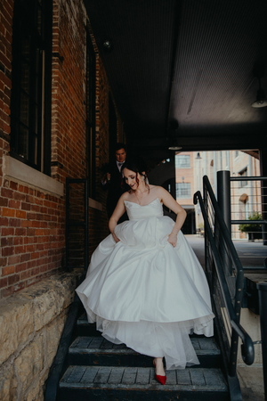 Bride in white ballgown with red heels walks up brick building steps, groom following behind during wedding photo session.
