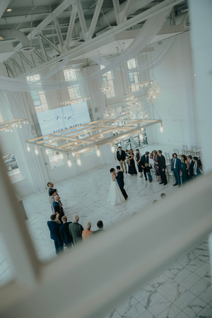 Aerial view of elegant white wedding reception venue with chandelier, guests gathered around couple on marble floor.