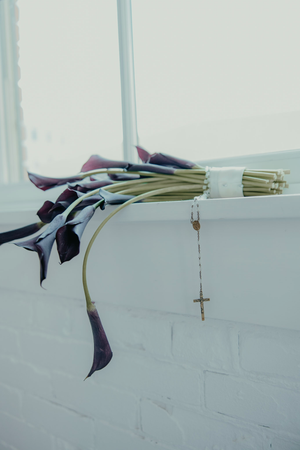 Dark purple calla lily bouquet with rosary beads resting on white brick ledge near window.