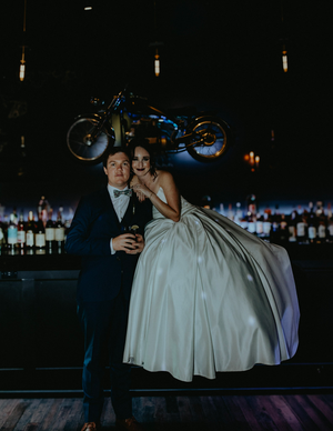 Bride in white ballgown sits on bar beside groom in navy suit, vintage motorcycle hanging above in dark reception venue.
