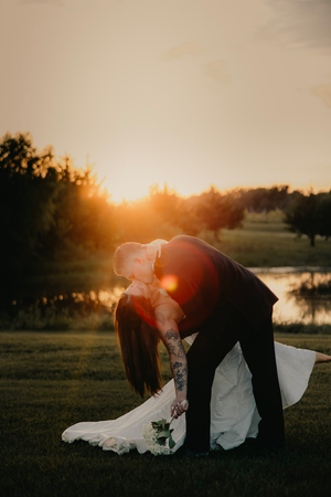 Bride and groom share a romantic dip kiss at golden hour sunset on lush green lawn.