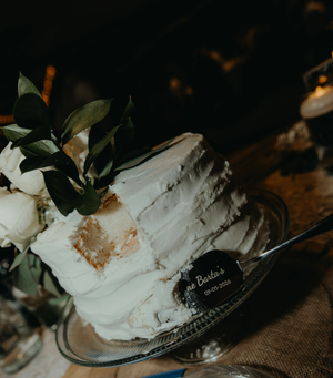 Elegant white rustic wedding cake with greenery and floral decor on glass stand.