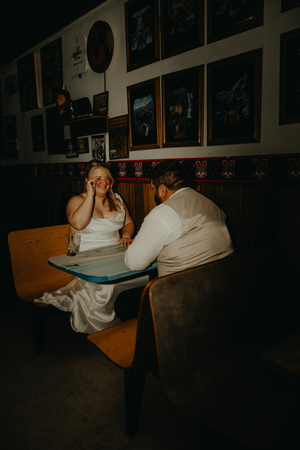 Bride wiping tears while groom leans in at rustic booth, framed art on walls, emotional wedding reception moment.