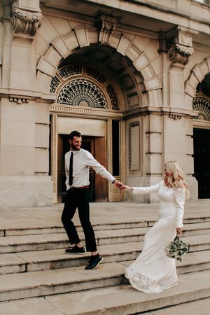 Bride and groom holding hands on stone steps outside an ornate building on their wedding day.