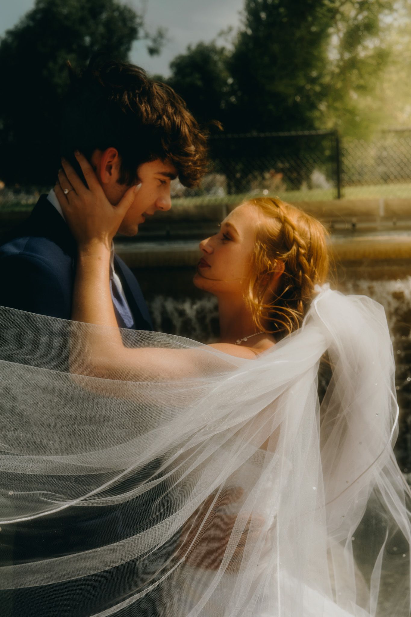 Bride and groom share a romantic moment, her veil flowing, near a sunlit pond.