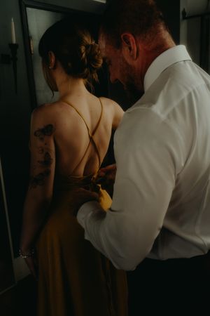 Man in white shirt fastening the back of a woman's golden backless dress with butterfly tattoo on her arm.