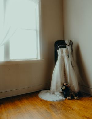 A white wedding dress with veil displayed on a hanger leaning against a chair near a sunlit window.