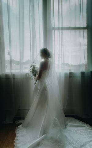 Bride in white wedding gown and veil holding bouquet, standing by sheer curtained window in soft natural light.