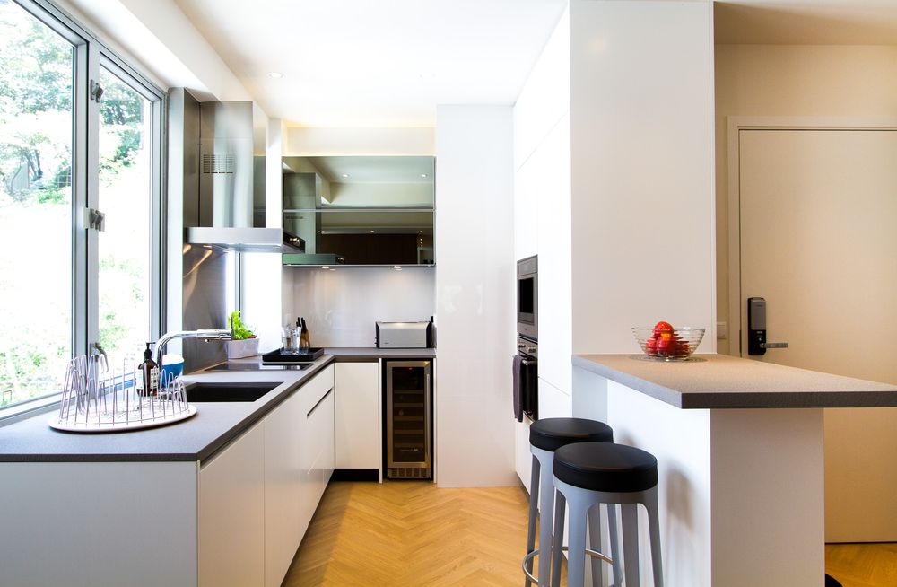 Modern white kitchen with herringbone parquet floor, island bar stools, and large window.