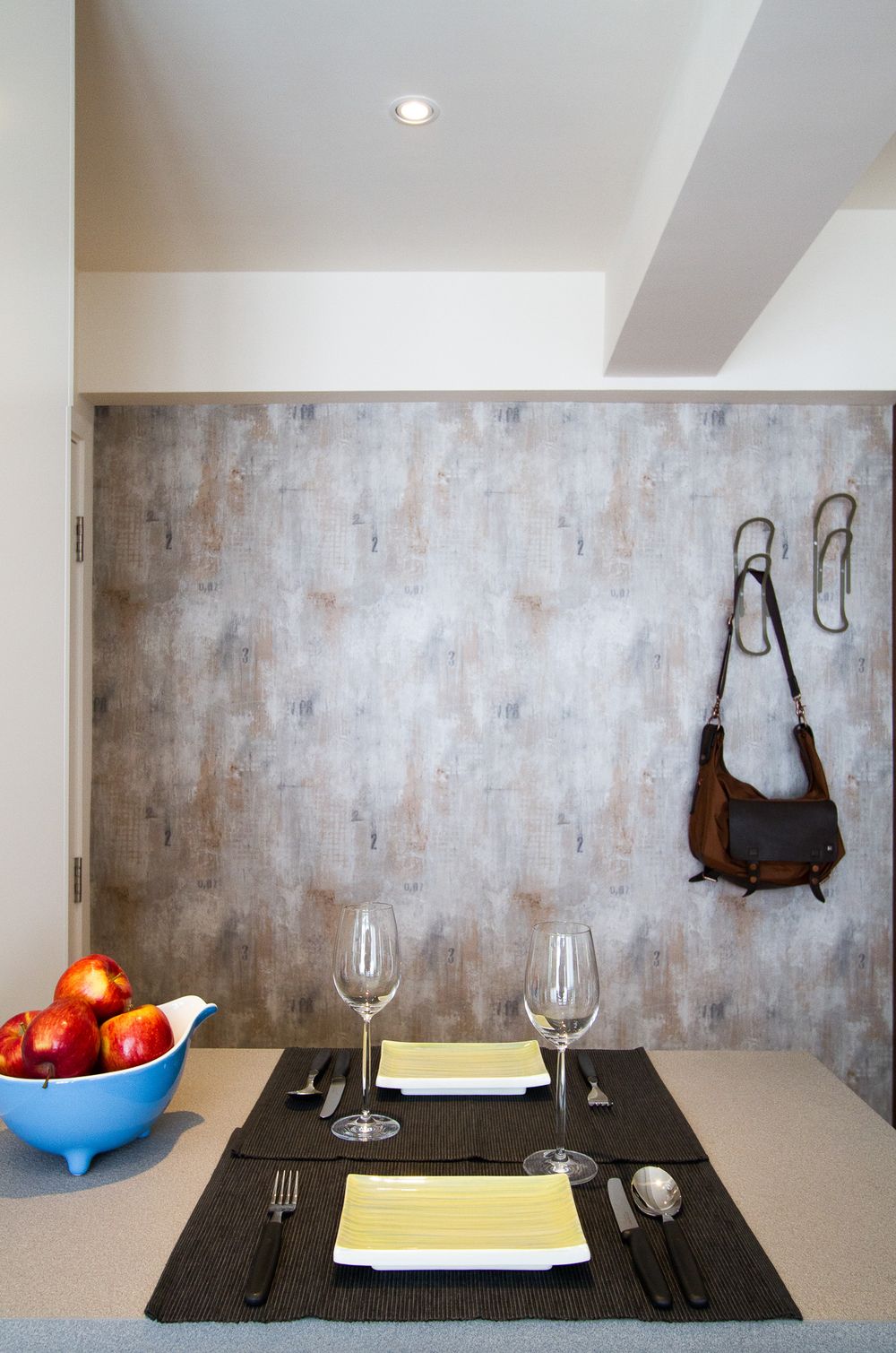Modern dining area with black placemats, wine glasses, and a blue bowl of apples against a textured concrete accent wall.